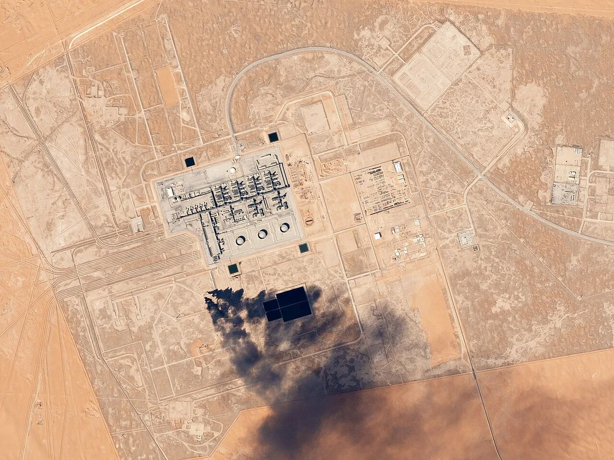 Aerial satellite image of an oil processing facility with tanks and infrastructure spread across desert terrain in Saudi Arabia.